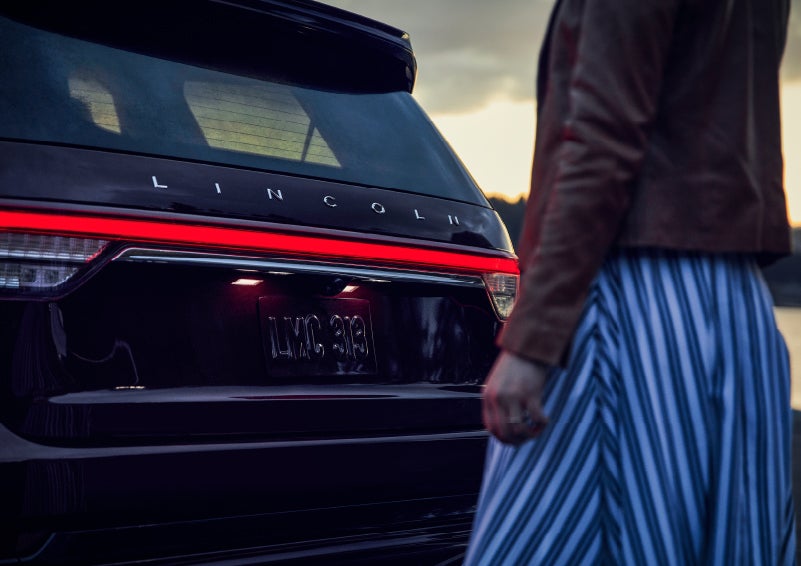 A person is shown near the rear of a 2024 Lincoln Aviator® SUV as the Lincoln Embrace illuminates the rear lights | Ed Morse Lincoln in Muscatine IA