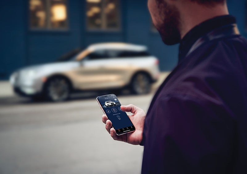 A person is shown interacting with a smartphone to connect to a Lincoln vehicle across the street. | Ed Morse Lincoln in Muscatine IA